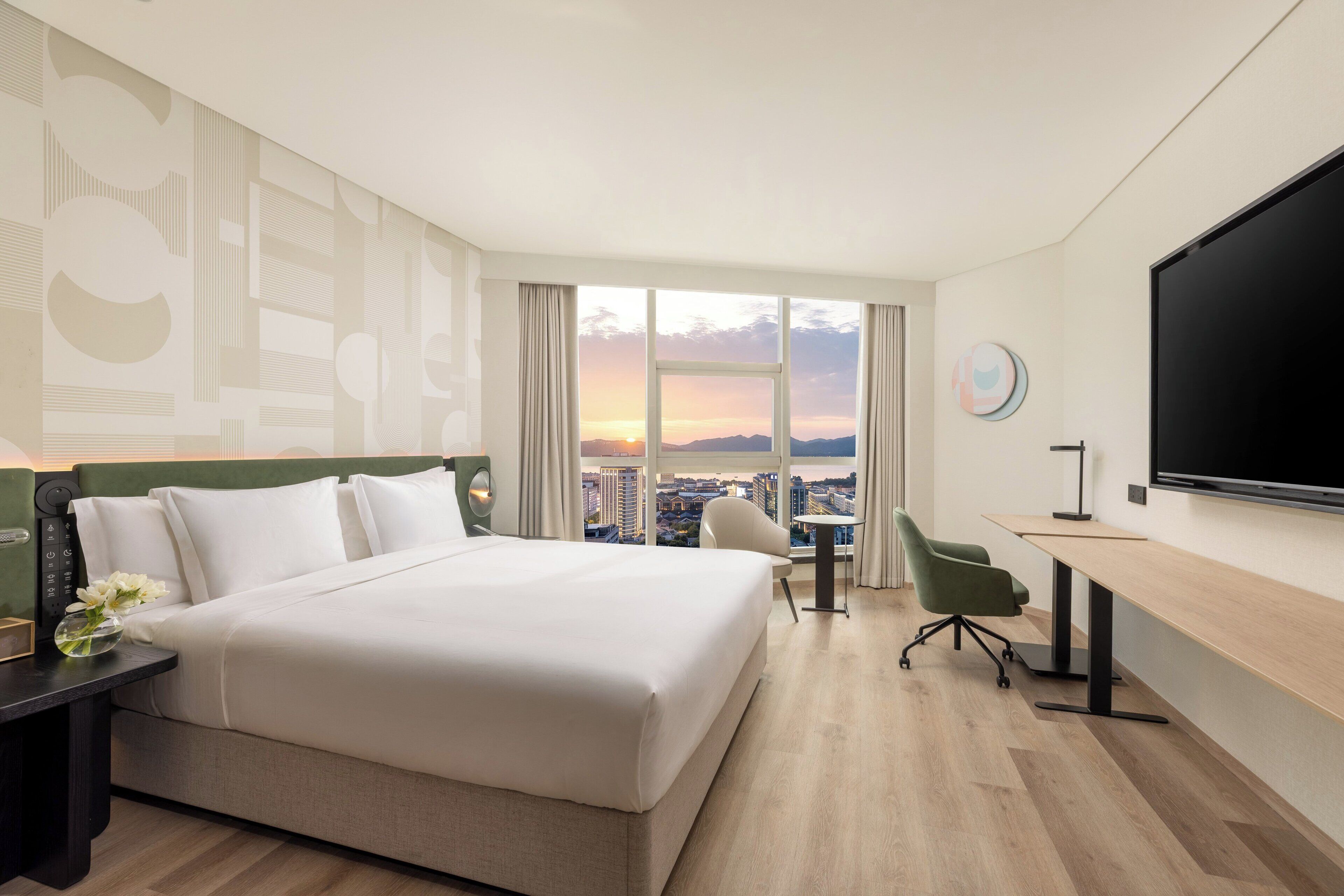 Habitación de hotel con una cama grande, un escritorio con una silla, un televisor y vistas a la ciudad al atardecer.