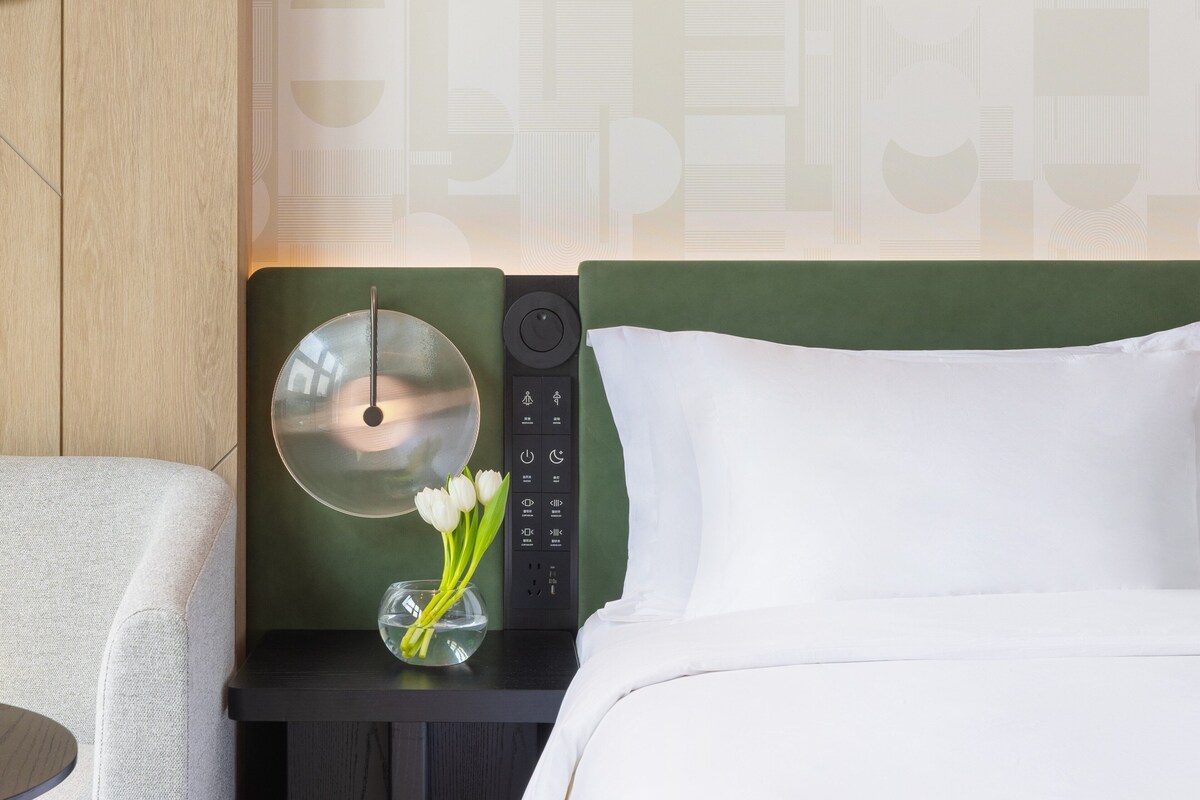 A hotel room with a bed, a bedside table with a lamp, a vase with flowers, and a control panel.