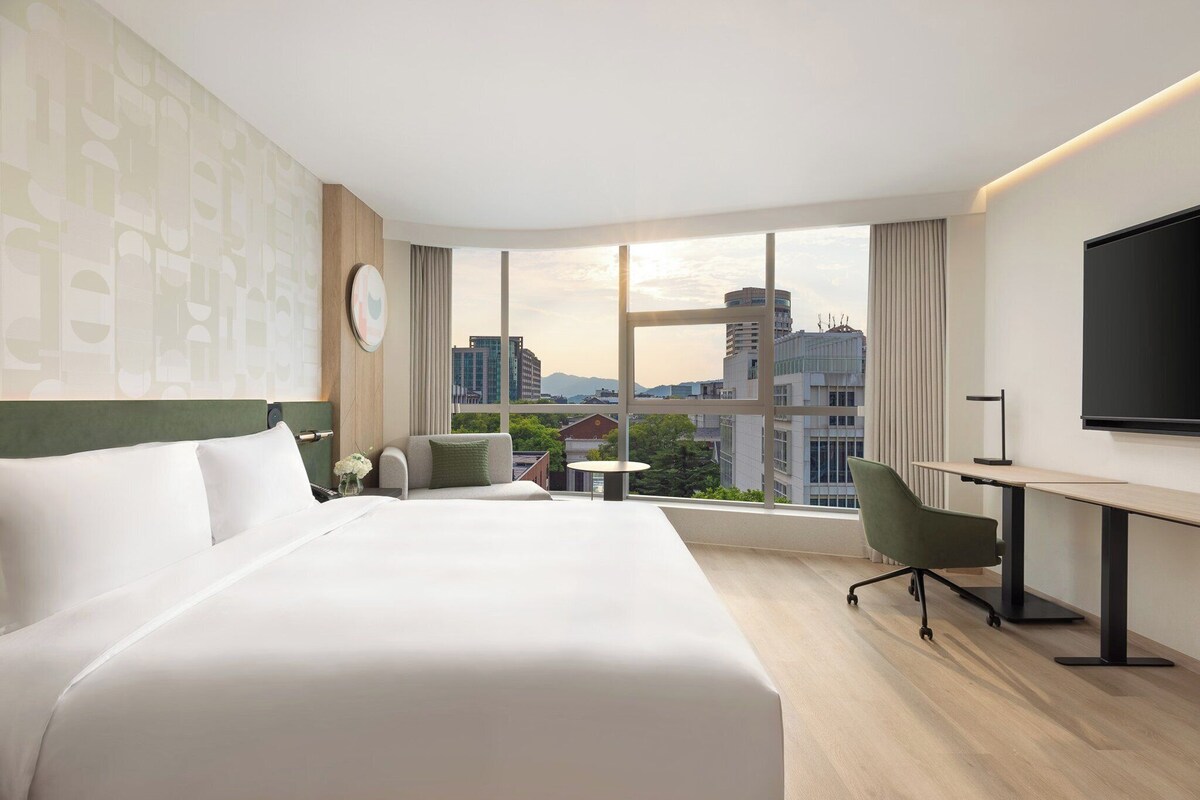 A hotel room with a large bed, a desk, a chair, a small table, and a TV.