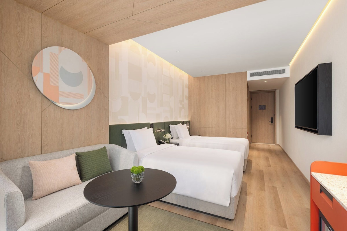 Una habitación de hotel moderna con dos camas, un sofá, una mesita con una planta y un televisor montado en la pared.