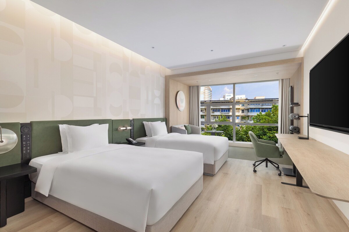 Une chambre d’hôtel moderne avec deux lits, un bureau et une grande fenêtre donnant sur la ville.