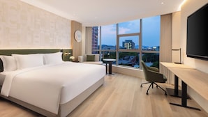 A modern hotel room with a large bed, a desk, and a view of the cityscape through large windows.