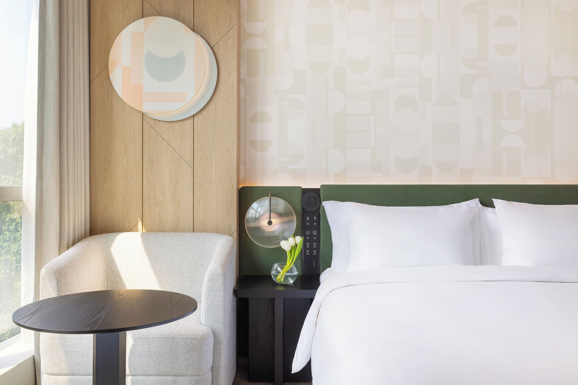 Une chambre d’hôtel comprenant un lit, une chaise, une petite table, un miroir fixé au mur et une table de chevet avec une télécommande.