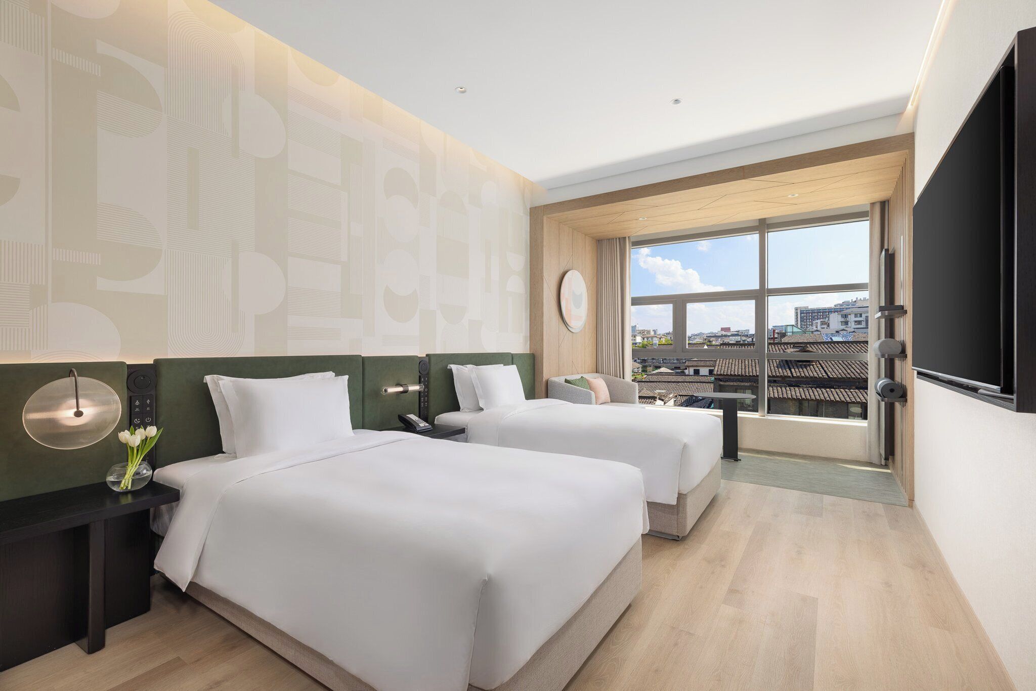 Una habitación de hotel moderna con dos camas, un ventanal amplio con vistas a la ciudad, un televisor de pantalla plana y una mesita de noche con un florero.
