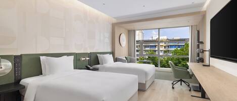 Habitación de hotel con dos camas, un escritorio y un amplio ventanal con vistas a edificios y vegetación.