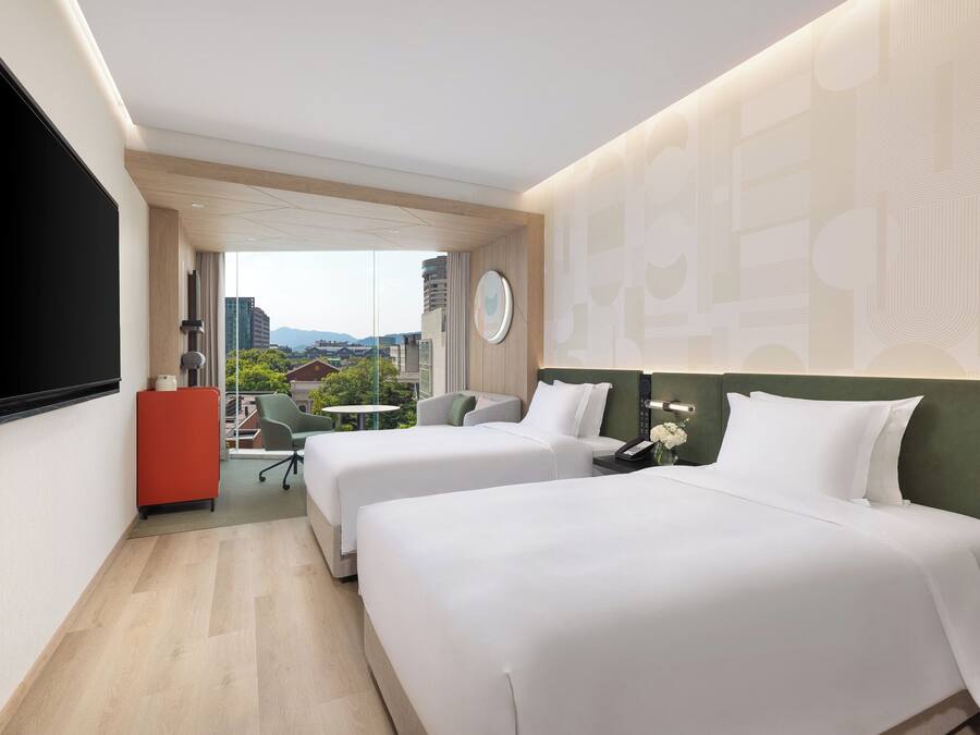 Habitación de hotel con dos camas, un escritorio, una silla y vistas a la ciudad.