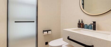 A modern bathroom with a glass shower door, a toilet, a sink with soap dispensers, and a mirror with a light.