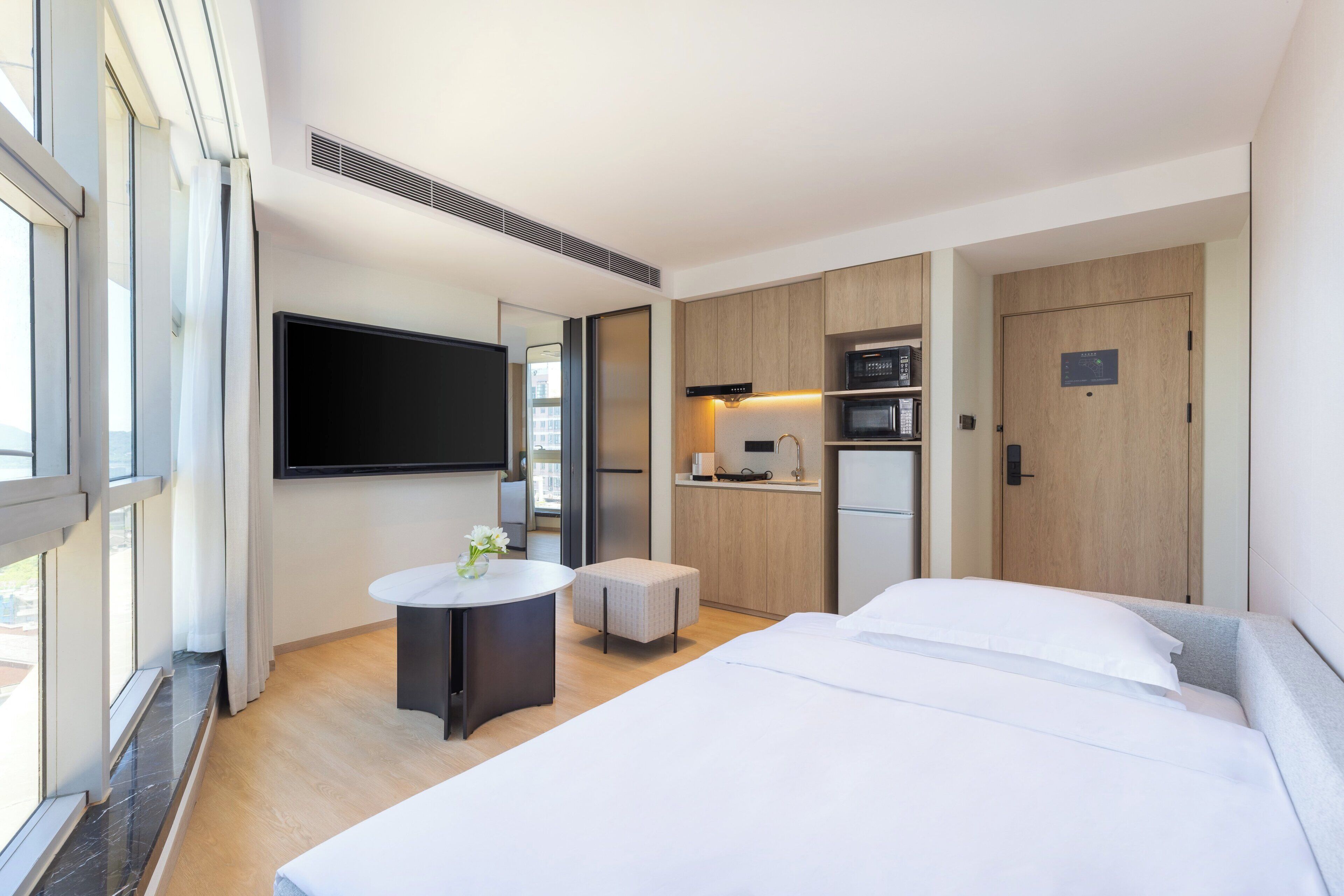A modern hotel room with a bed, a small round table with a vase, a kitchenette, and a TV mounted on the wall.