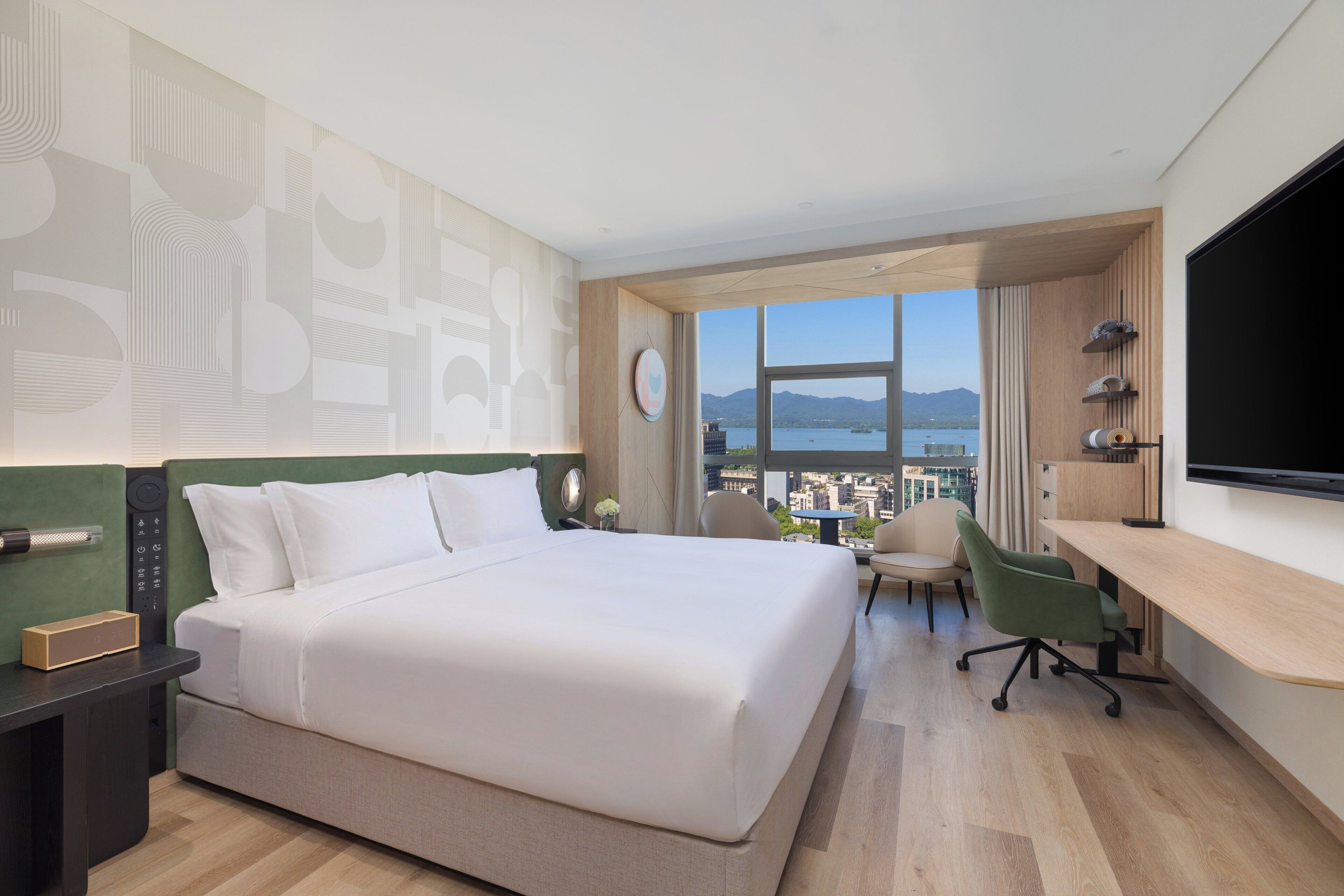 Habitación de hotel con una cama grande, un escritorio, una silla, un televisor y vistas a un cuerpo de agua y montañas.