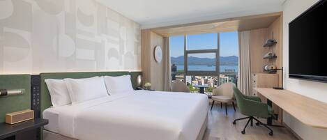Habitación de hotel con una cama grande, un escritorio, una silla, un televisor y vistas a un cuerpo de agua y montañas.