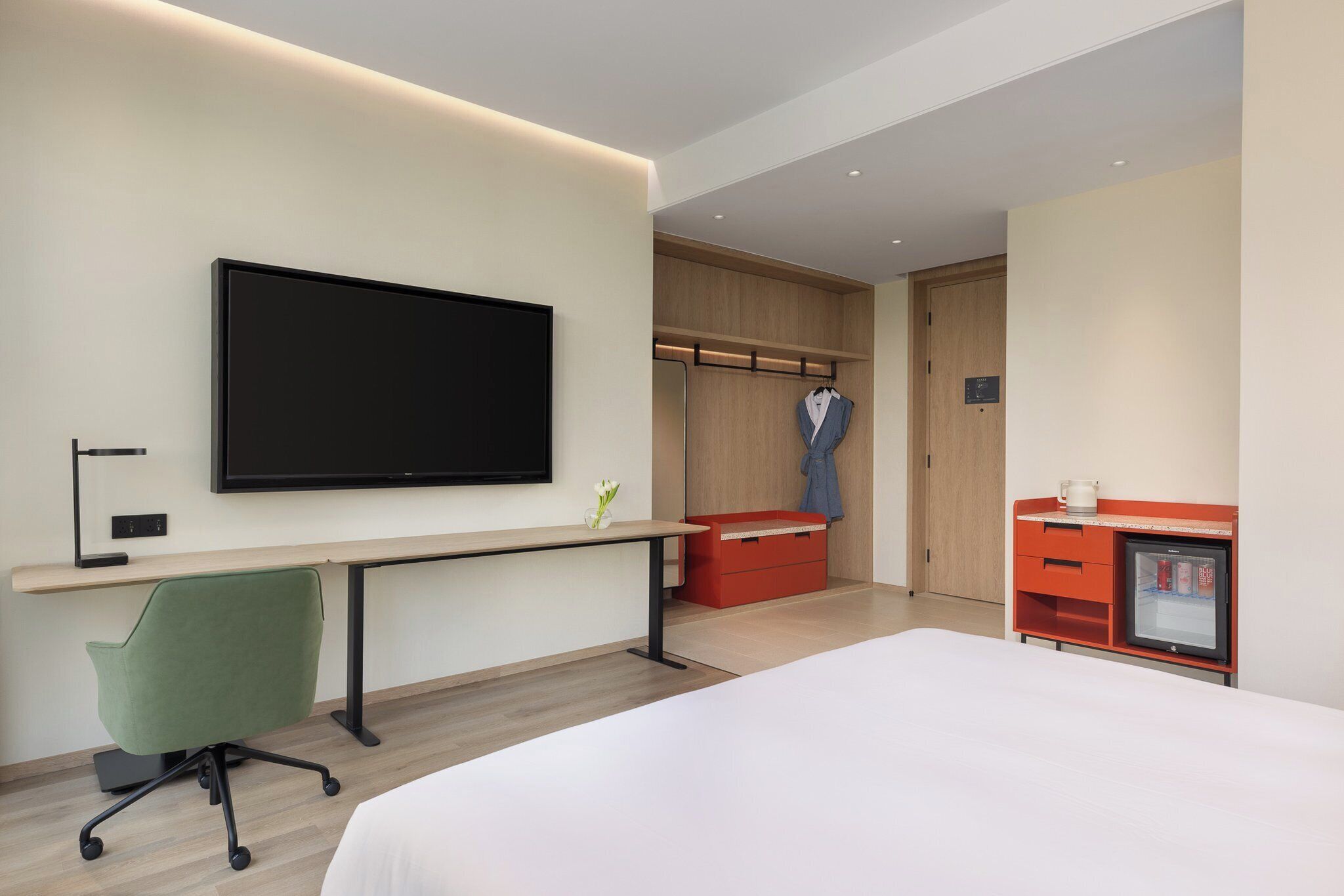 A modern hotel room with a large bed, a desk with a computer, a flat-screen TV, a wardrobe, and a minibar.