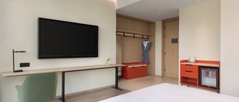 A modern hotel room with a large bed, a desk with a computer, a flat-screen TV, a wardrobe, and a minibar.