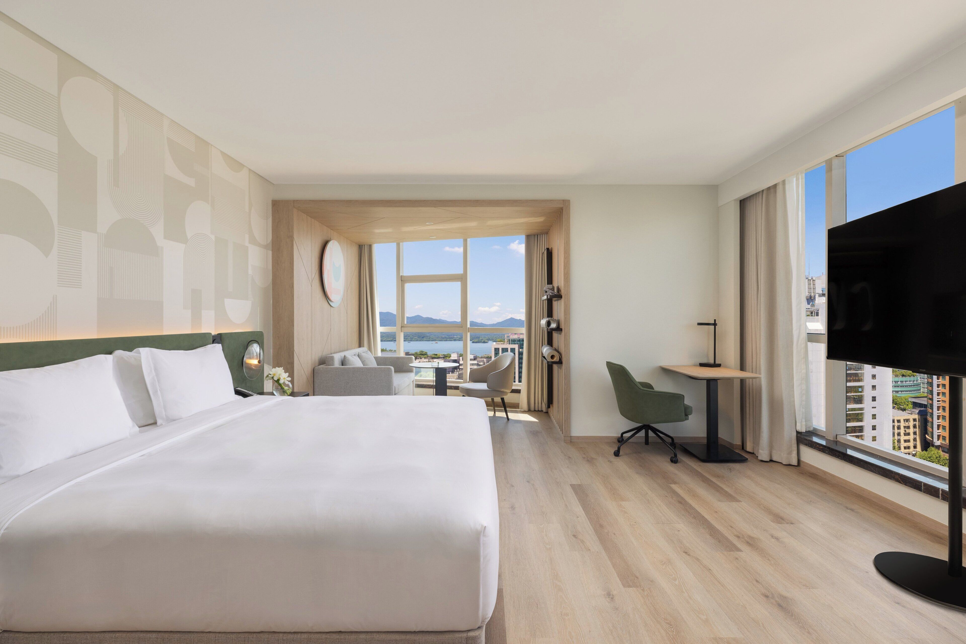 Habitación de hotel con una cama grande, un escritorio, una silla y un televisor. Hay una ventana que ofrece vistas a edificios y montañas.