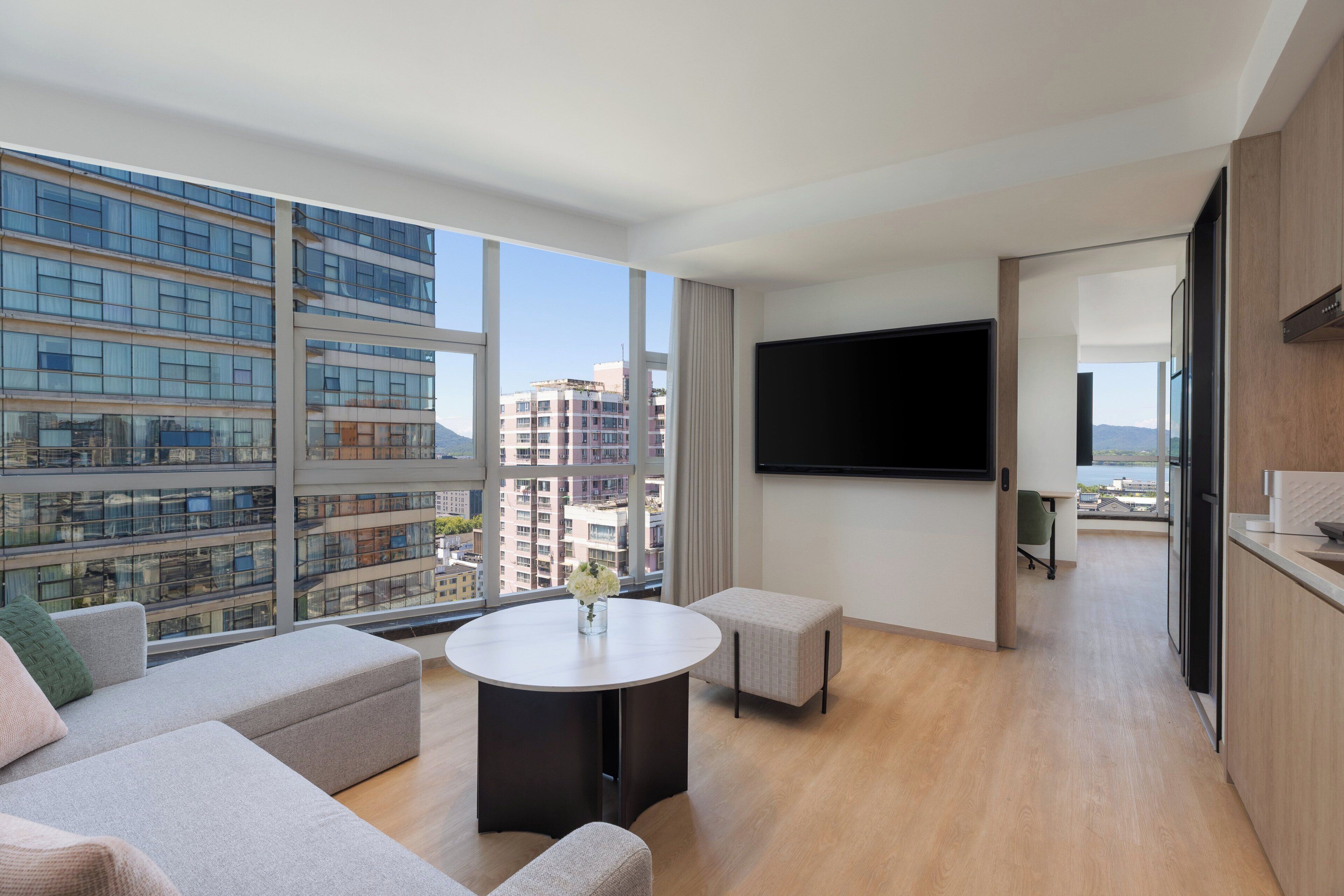 A modern hotel room with a large window, a flat-screen TV, a round coffee table, and a sofa.