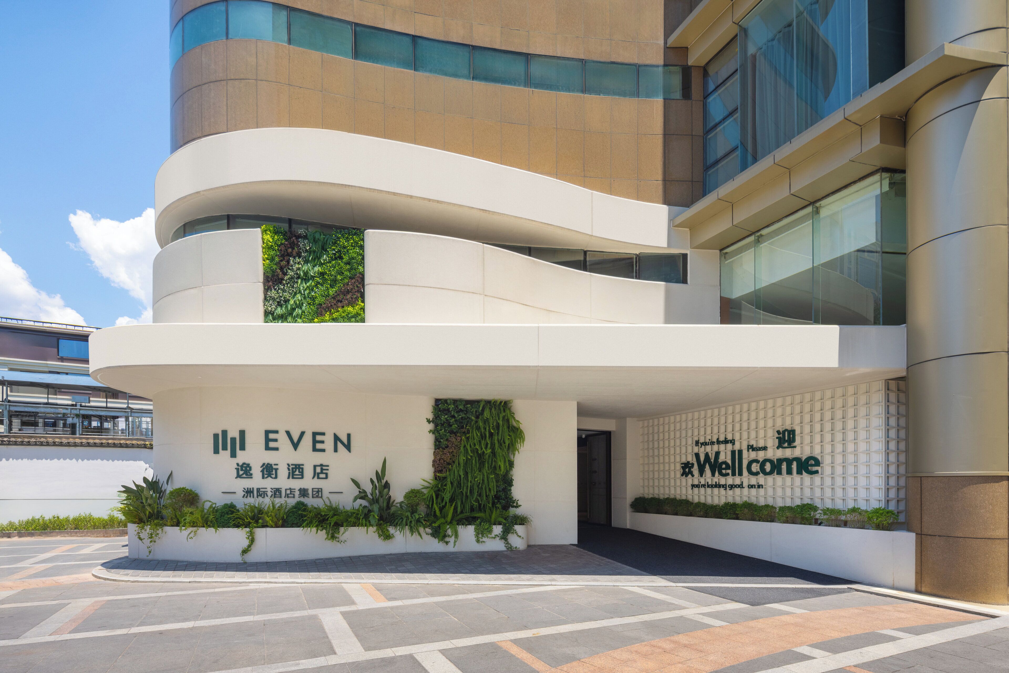 Una entrada de hotel moderna con fachada curva, un letrero de bienvenida y un área ajardinada con plantas.