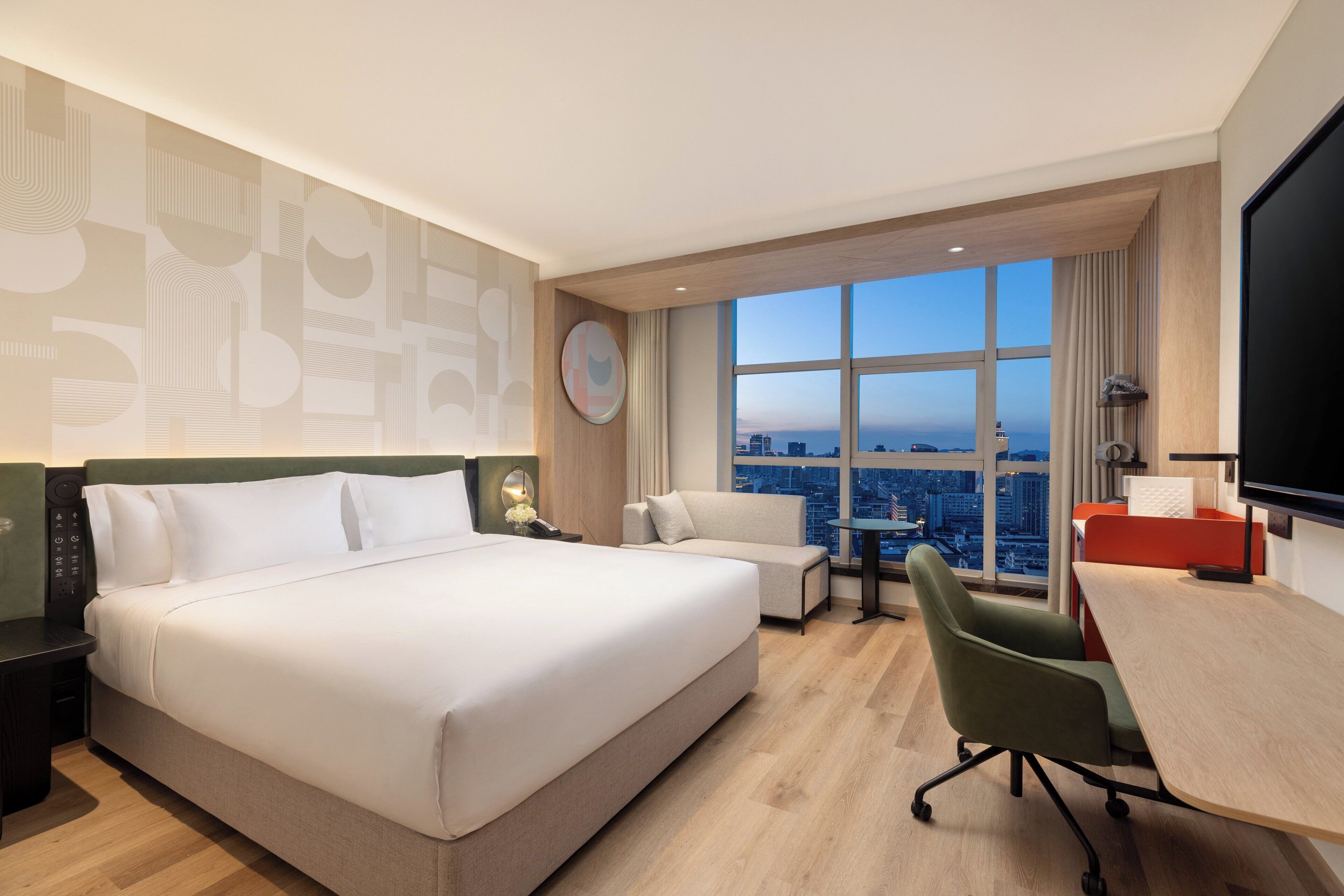 A hotel room with a large bed, a desk, a chair, and a view of the cityscape through large windows.
