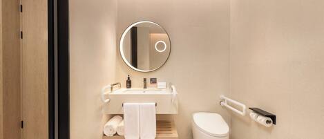 A modern bathroom with a wooden door, a round mirror, a sink with toiletries, a toilet, and a towel rack.
