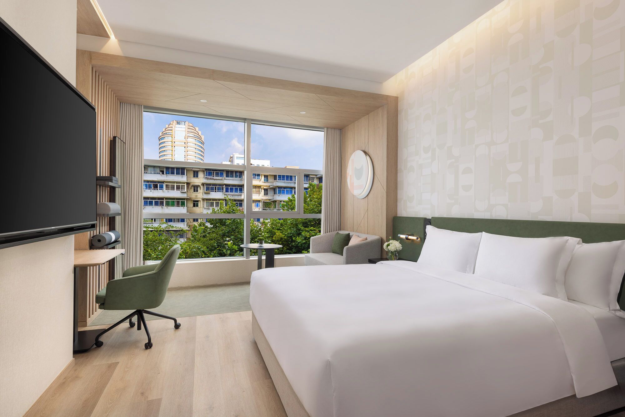 A hotel room with a large bed, a desk, a chair, a TV, and a view of a building through the window.