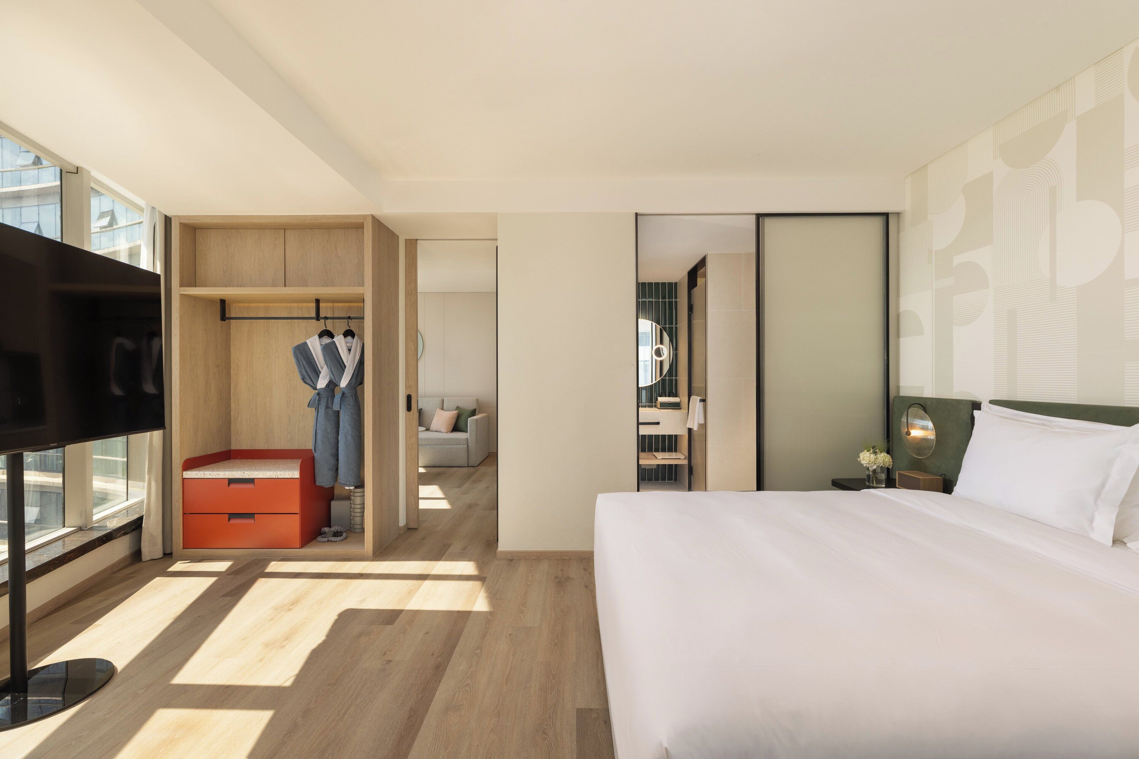 A modern hotel room with a large bed, a TV, a wardrobe, and a bathroom visible through an open door.