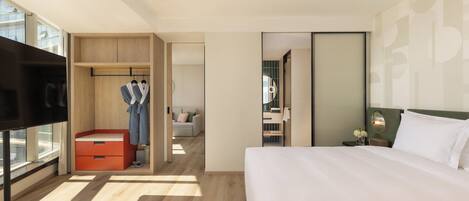 A modern hotel room with a large bed, a TV, a wardrobe, and a bathroom visible through an open door.