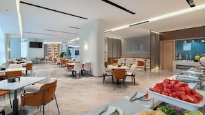 A modern hotel dining area with multiple tables and chairs, a buffet with various fruits, and a television mounted on the wall.