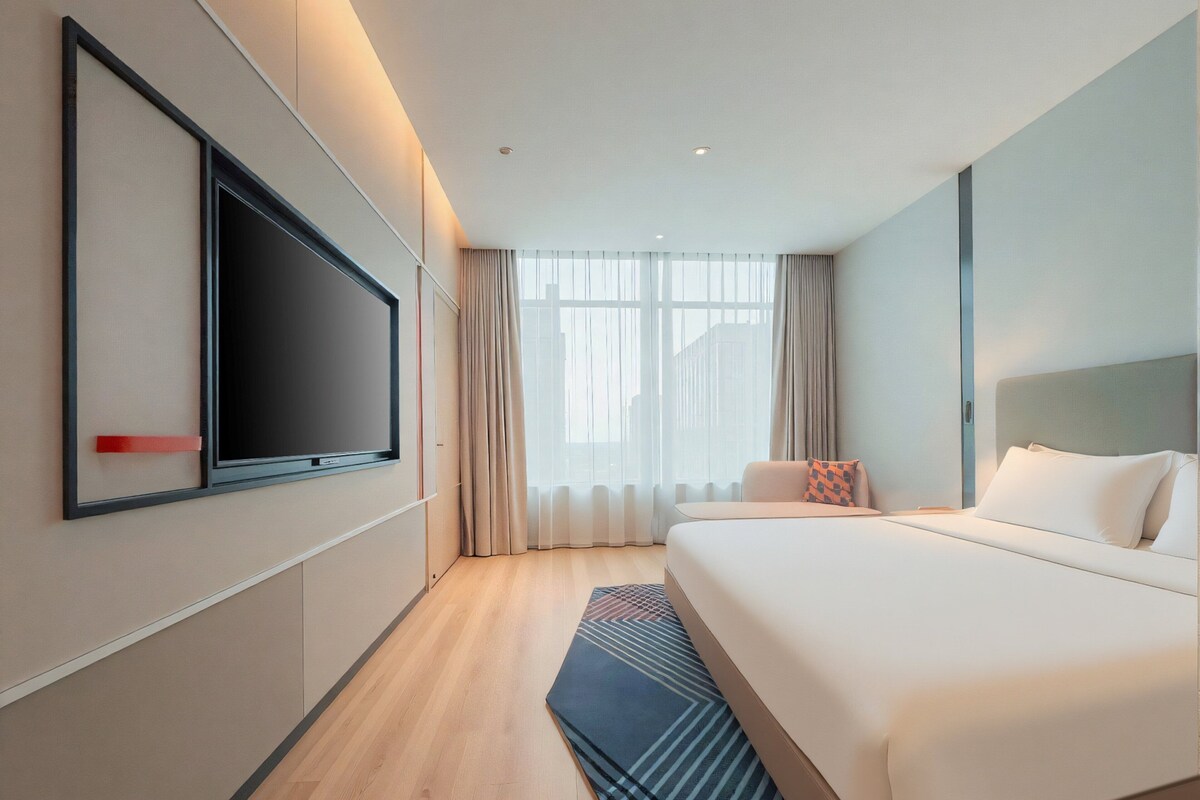 A hotel room with a large bed, a flat-screen TV mounted on the wall, and a view of the city through large windows.