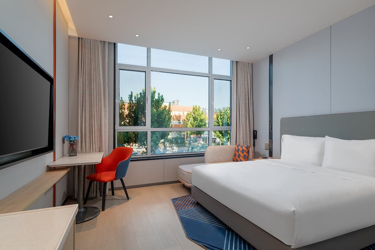 Quarto de hotel moderno com uma cama grande, uma mesa de escritório, uma cadeira e vista da cidade através de grandes janelas.