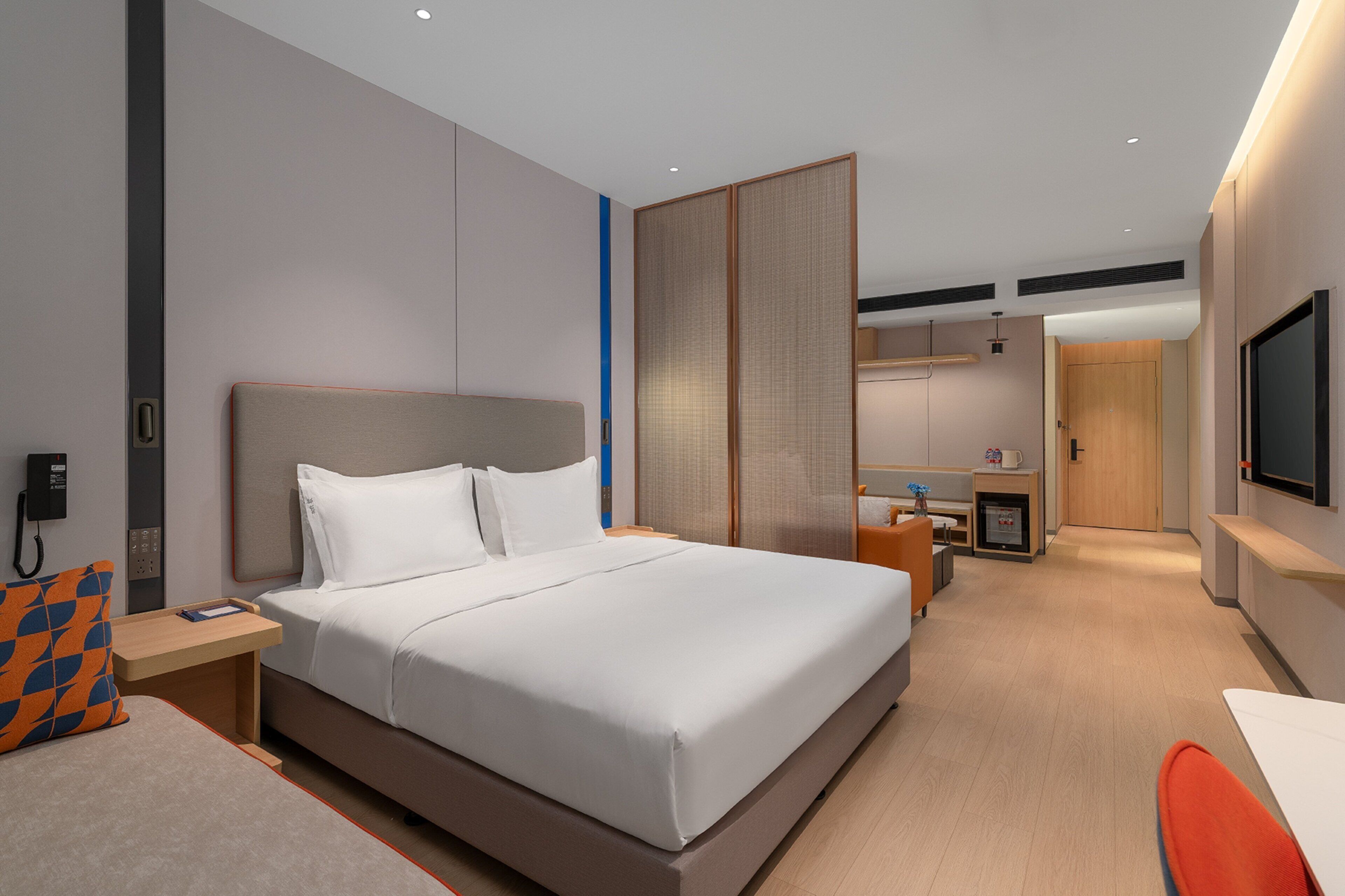 A modern hotel room with a large bed, a desk, a chair, a TV, and a bathroom visible in the background.