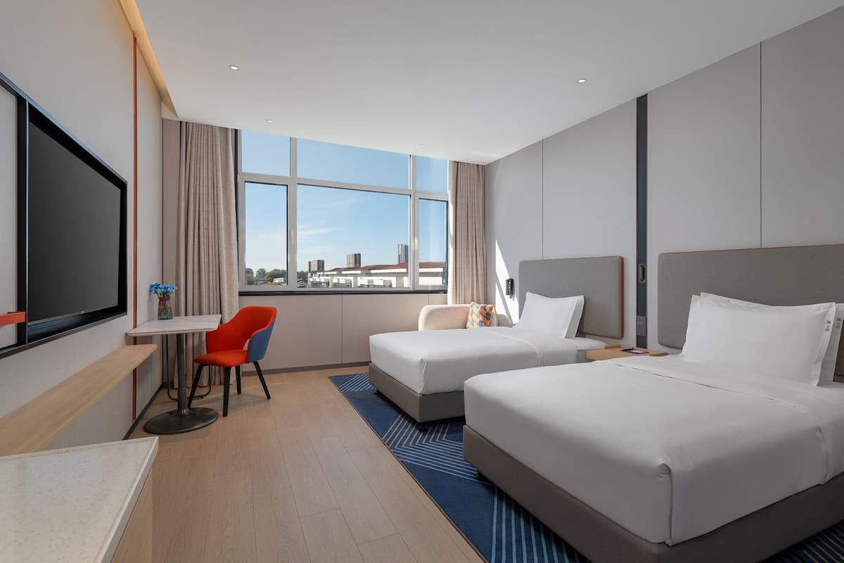 Quarto de hotel com duas camas, uma escrivaninha, uma cadeira e uma janela grande com vista para a cidade.