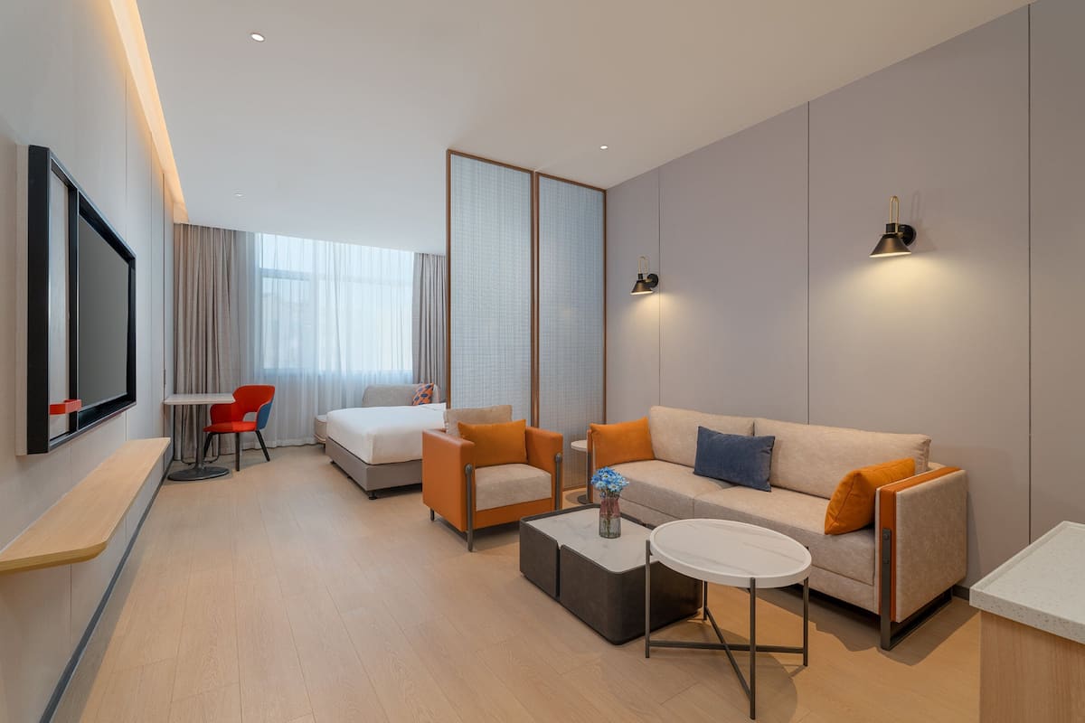 Quarto de hotel moderno com sofá, poltronas, mesa de centro, televisão e janela com cortinas.
