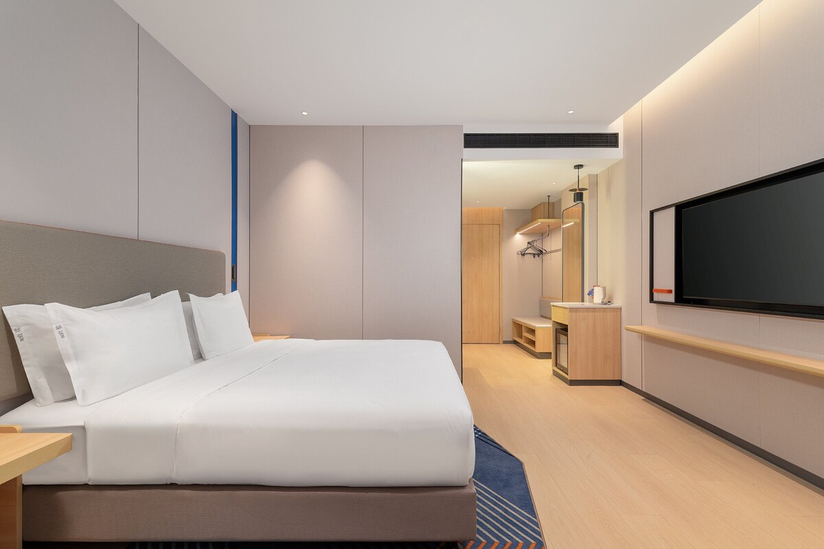 A modern hotel room with a large bed, a flat-screen TV mounted on the wall, a small desk, and a wooden nightstand.