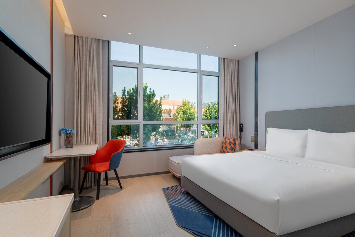 A modern hotel room with a large bed, a desk, a chair, and a view of the city through large windows.