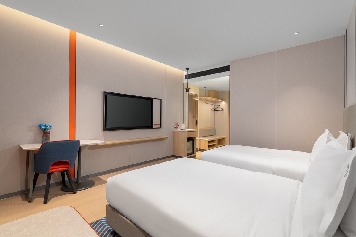 A hotel room with two beds, a desk with a chair, a TV mounted on the wall, and a mirror above the desk.
