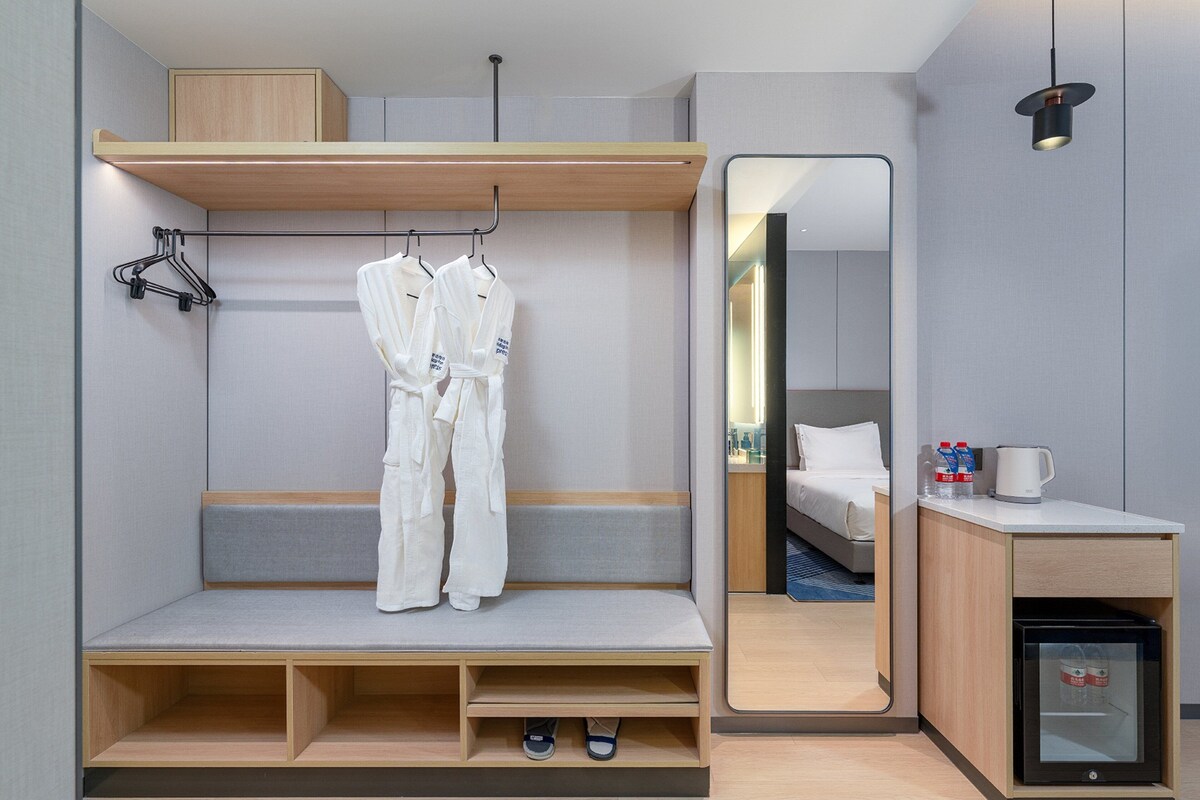 A modern hotel room with a built-in wardrobe, a mirror, a small kitchenette, and a seating area.