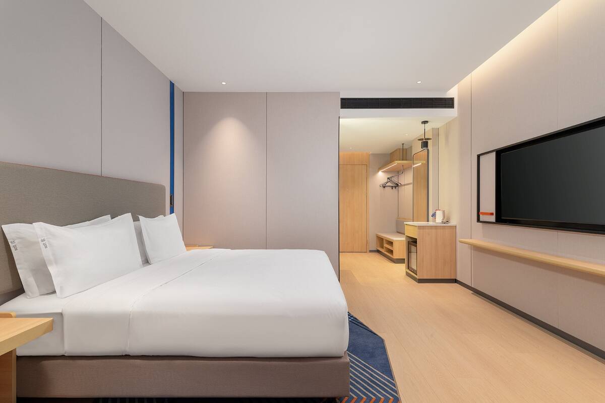 Quarto de hotel moderno com uma cama grande, televisão de tela plana na parede, uma pequena escrivaninha e uma mesa de cabeceira de madeira.