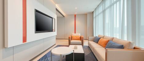 Una habitación de hotel moderna con un televisor de pantalla plana grande montado en la pared, un sofá con cojines naranjas, una mesita redonda pequeña y una alfombra estampada.