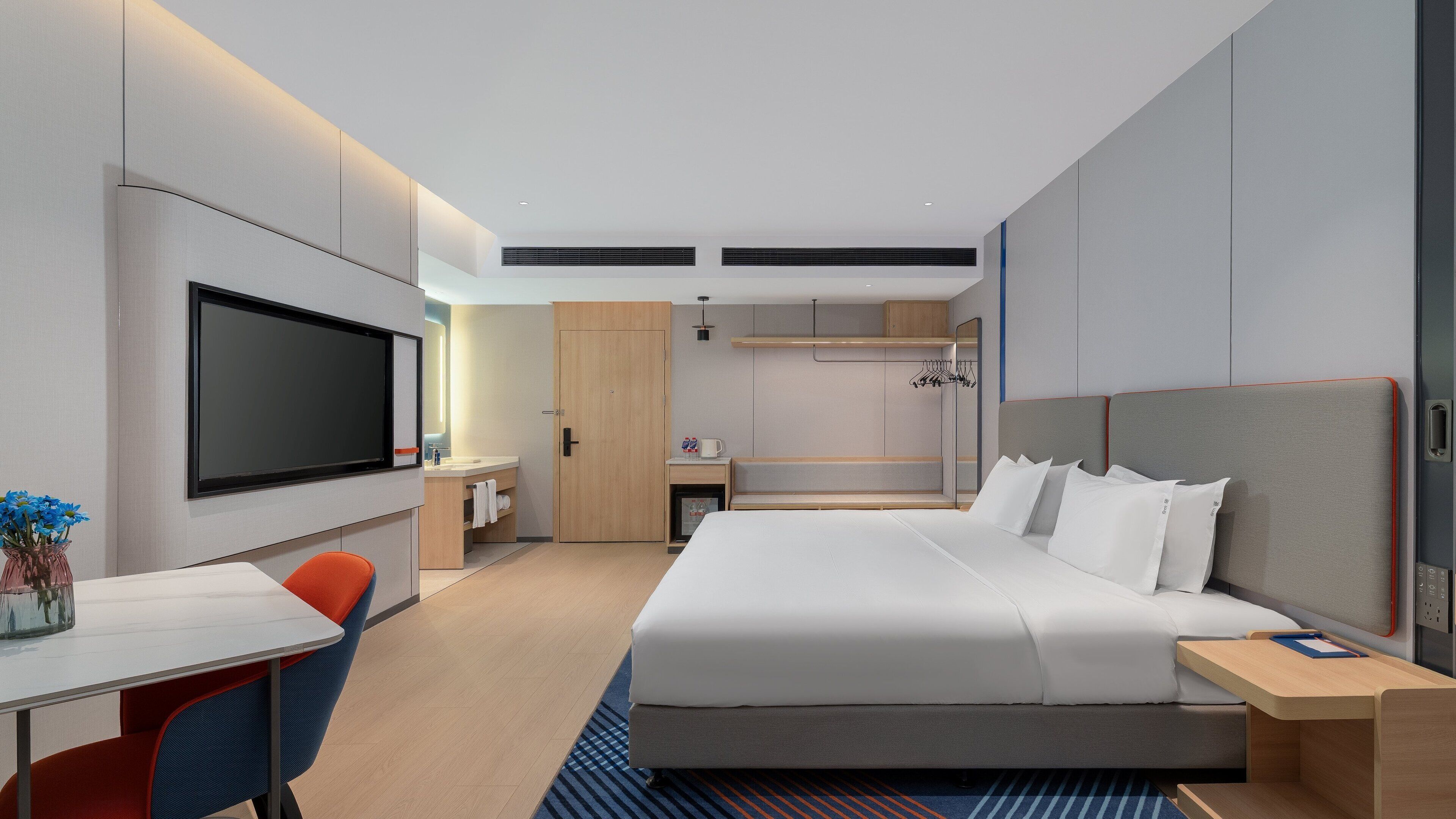 A modern hotel room with a large bed, a desk, a flat-screen TV, a wardrobe, and a small kitchenette.