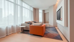 A modern hotel room with a large window, a sofa, a small round table, and a flat-screen TV mounted on the wall.