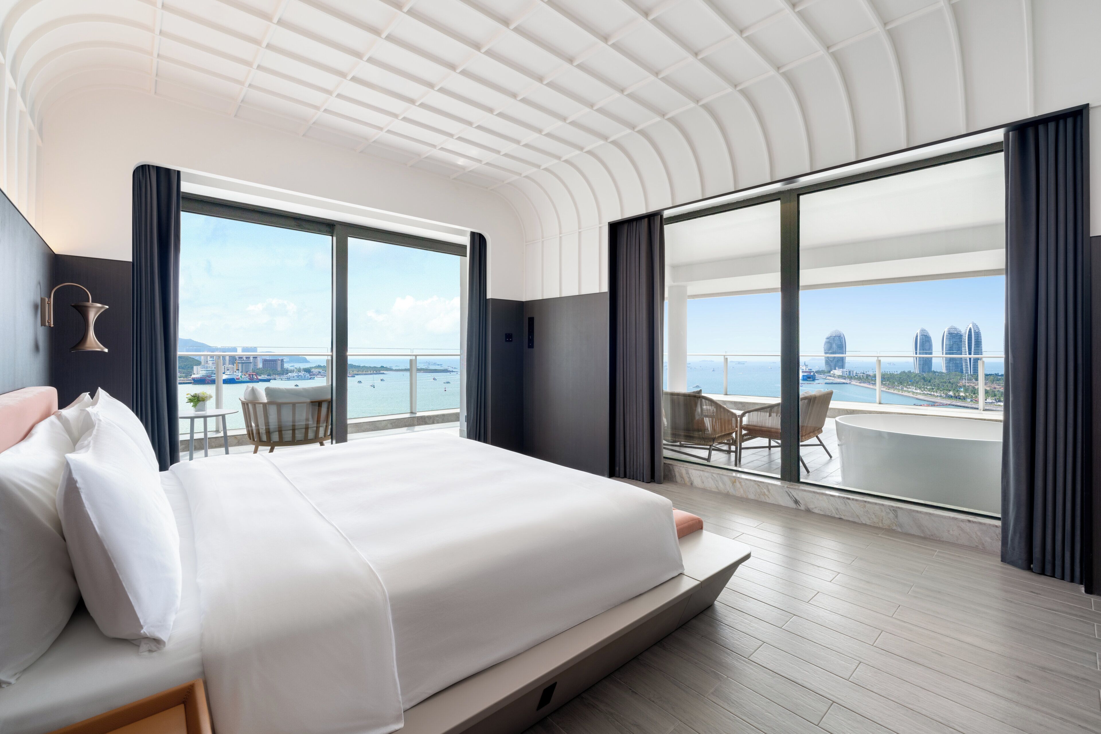 A modern hotel room with a large bed, a balcony with a view of the city, and a table with chairs.