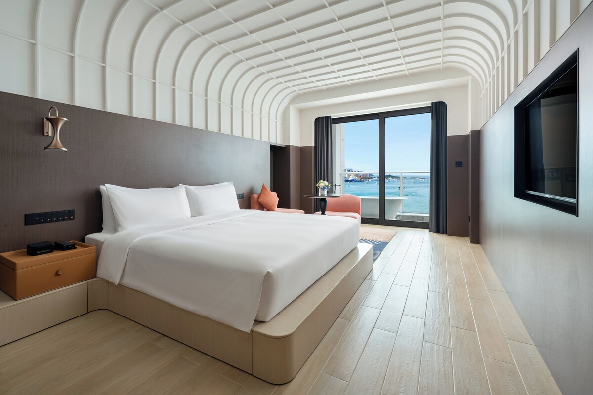 Habitación de hotel moderna con una cama grande, una mesita de noche, un televisor y vistas al mar.