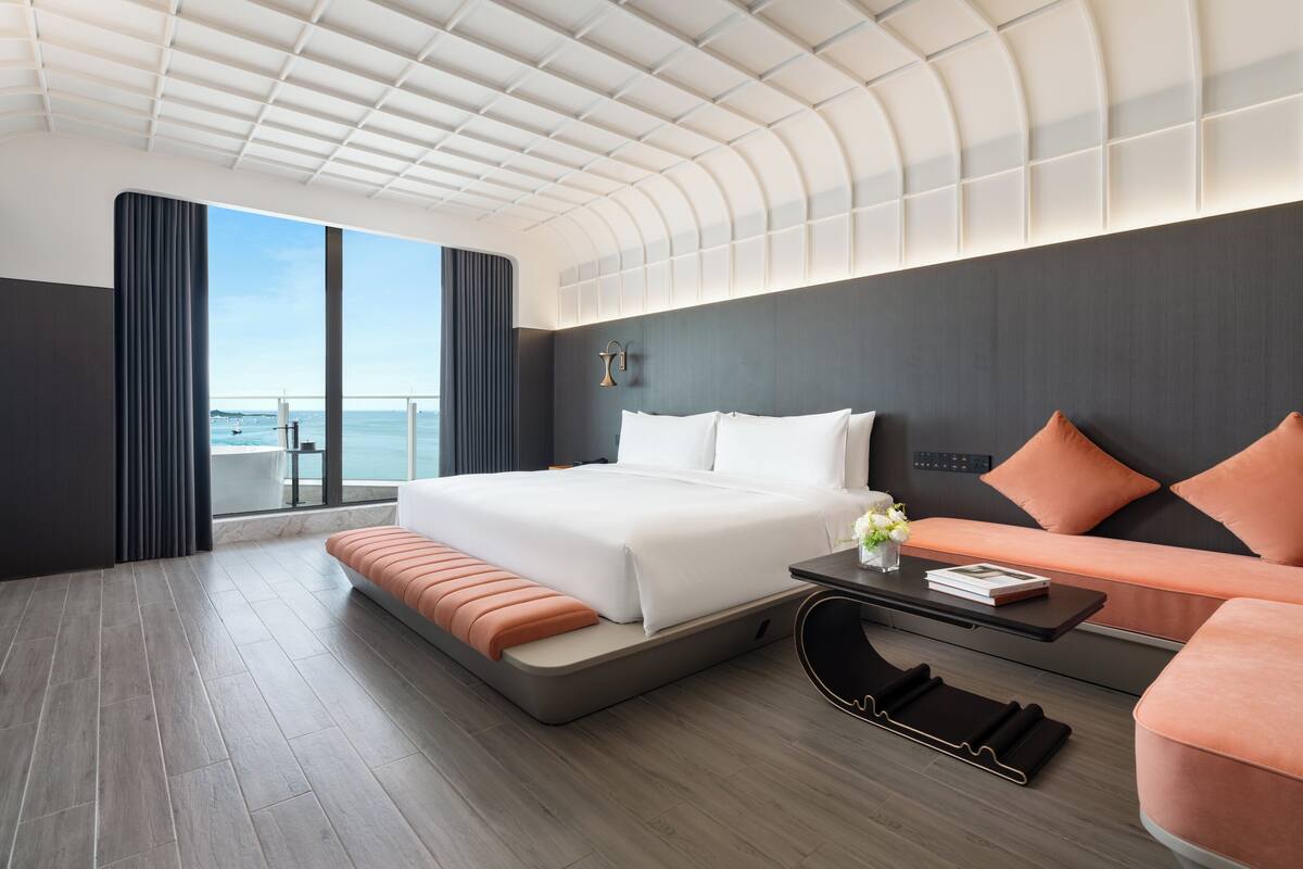 A modern hotel room with a large bed, a sofa, a small table, and a view of the sea.