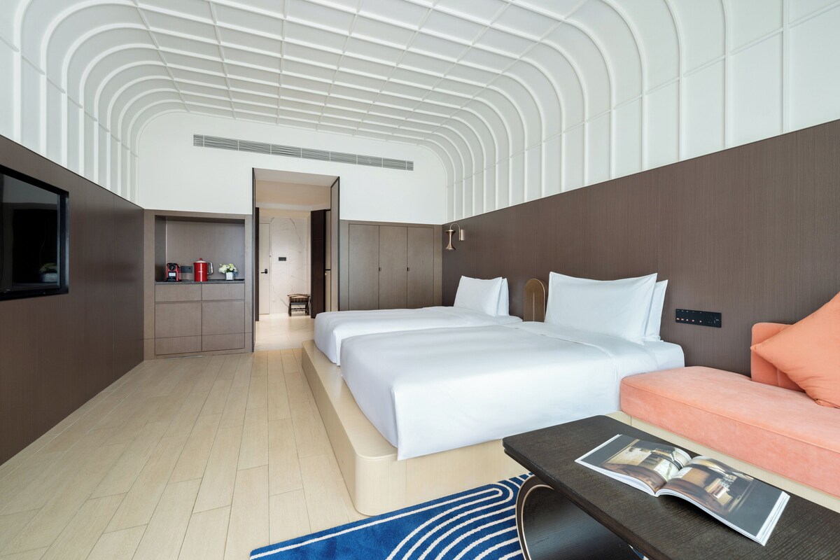 A hotel room with a modern design, featuring two beds, a sofa, a flat-screen TV, and a small kitchenette.