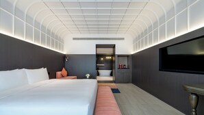Una habitación de hotel moderna con una cama grande, un televisor de pantalla plana, una pequeña zona de estar y un baño visible a través de una puerta abierta.