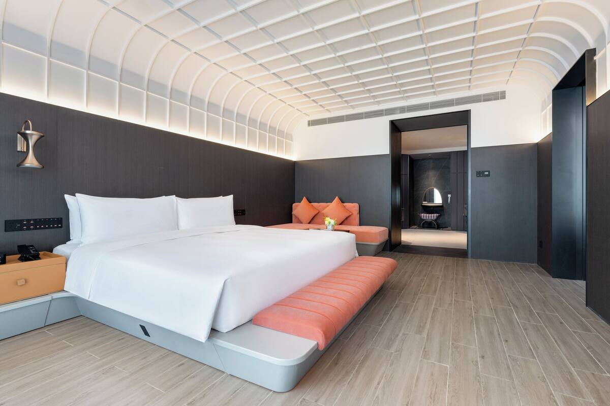 A modern hotel room with a large bed, a bench, a nightstand, a wall-mounted lamp, and a ceiling with a unique design.