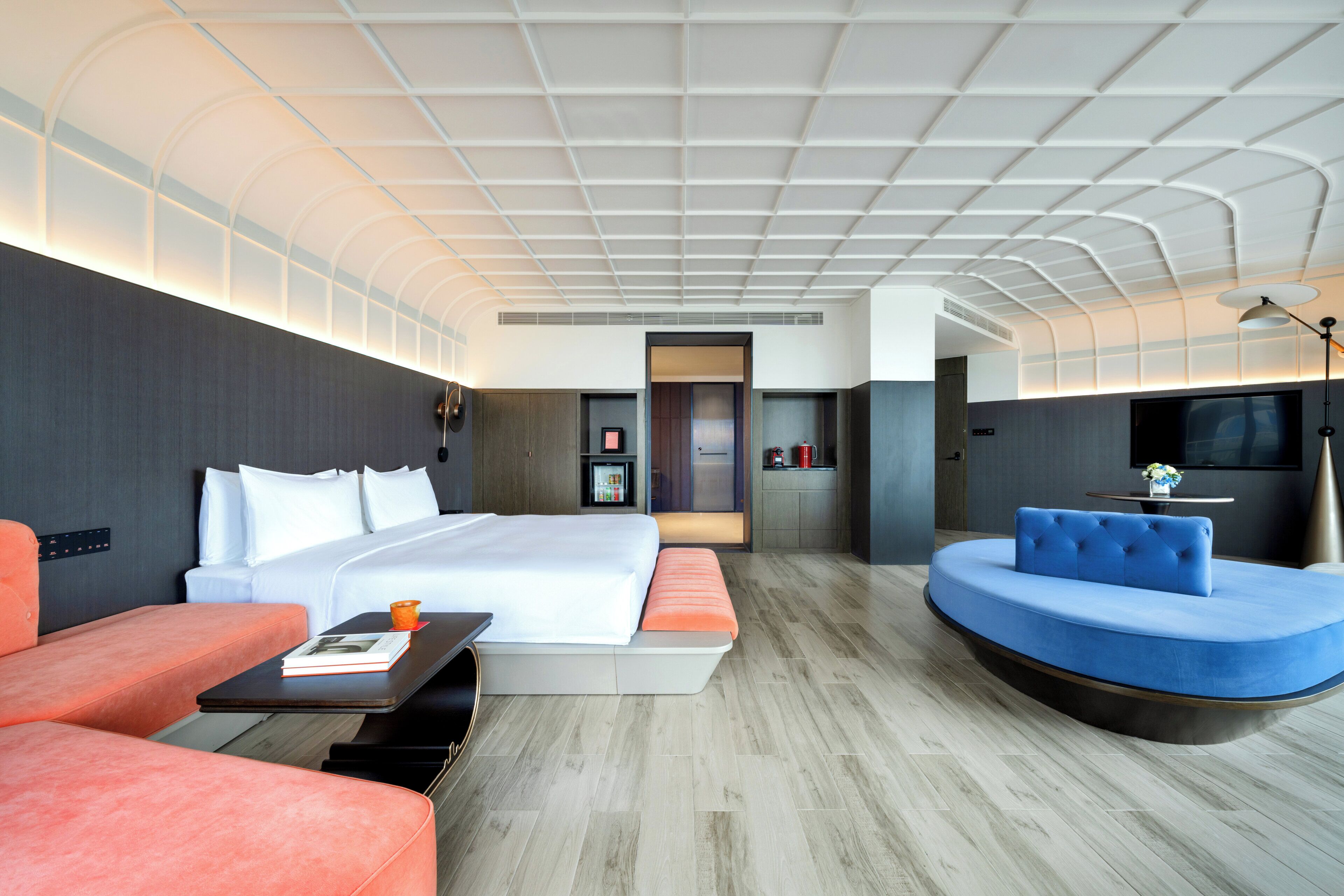 A modern hotel room with a large bed, a sofa, a curved seating area, a small kitchenette, and a TV.