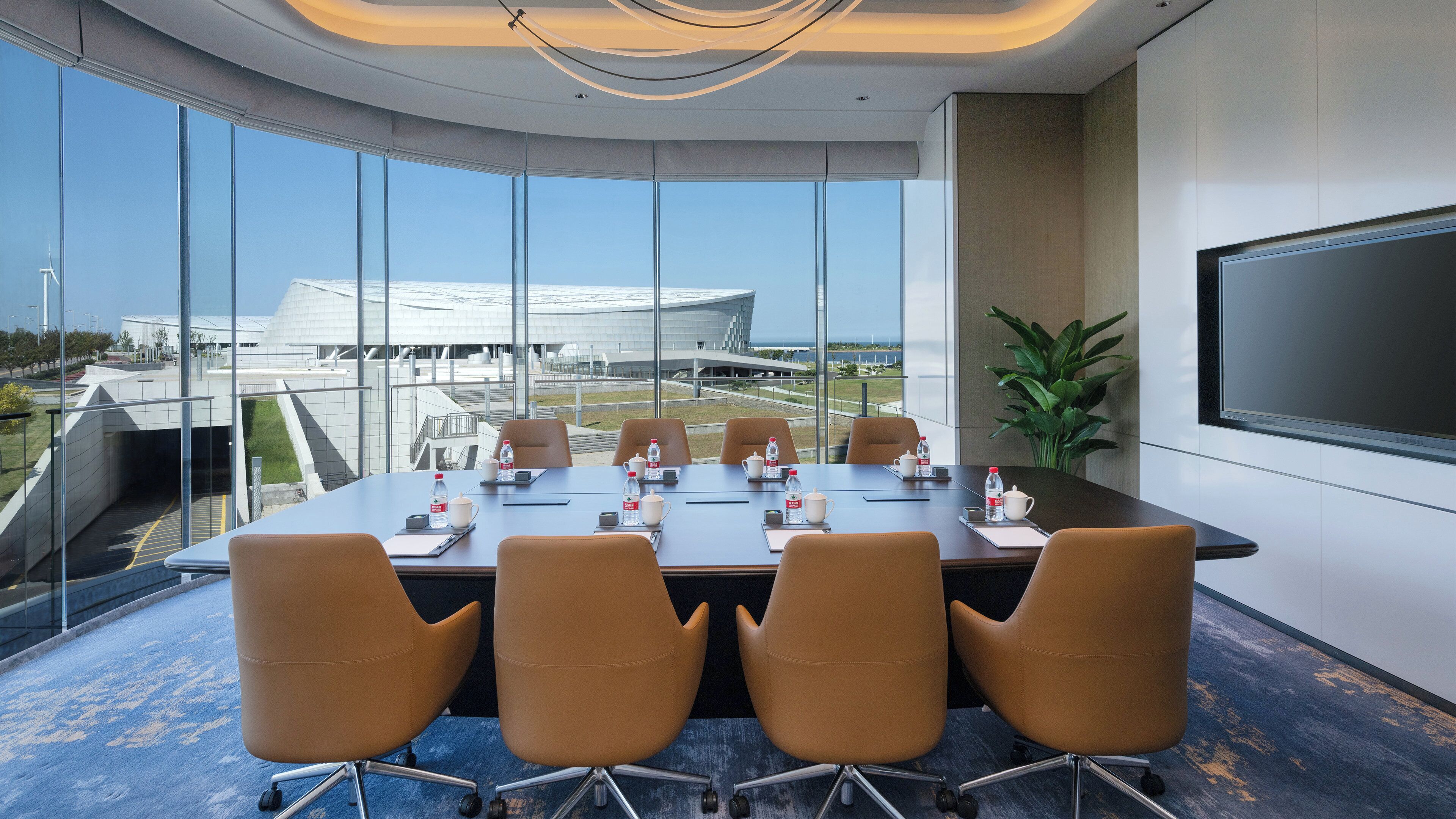 Un salón de conferencias con una gran mesa, cuatro sillas, una pantalla de televisión y vistas a un edificio moderno a través de amplios ventanales.