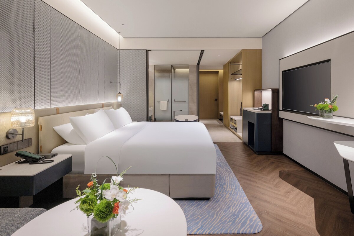 A modern hotel room with a large bed, a TV, a desk, and a bathroom visible through a glass door.