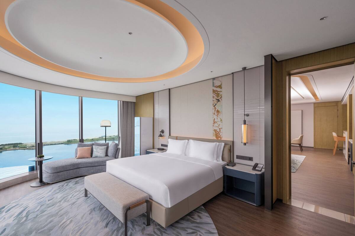 A modern hotel room with a large bed, a seating area with a view of the water, and a separate sitting area with a view of the interior.