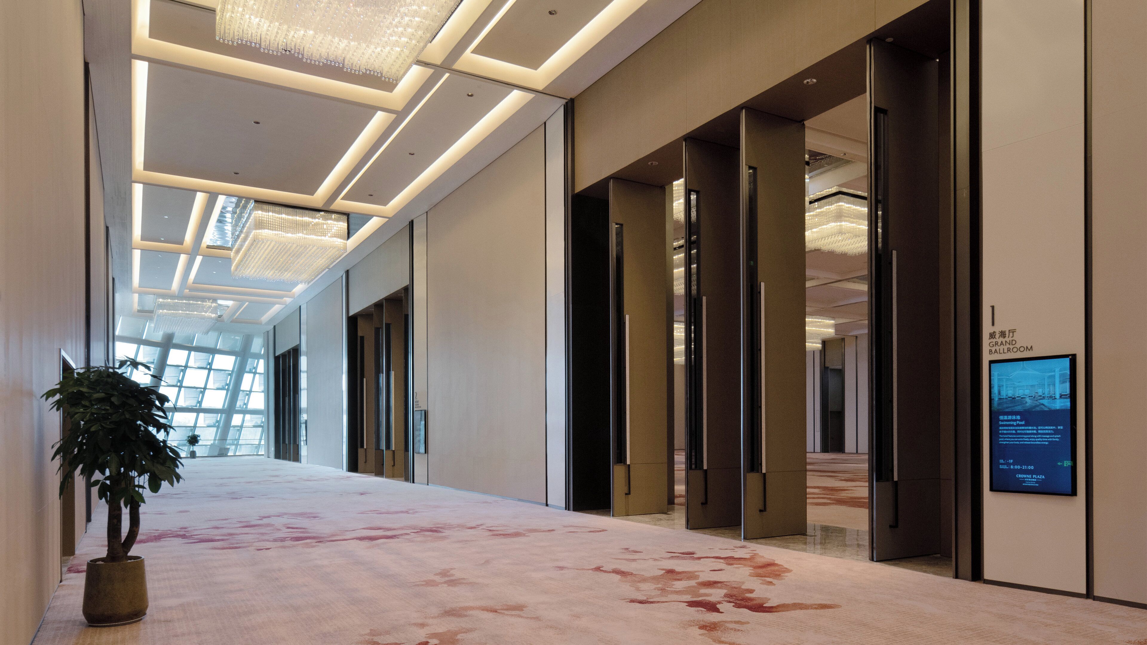 A modern hotel corridor with a carpeted floor, large windows, and a sign indicating the Grand Ballroom.