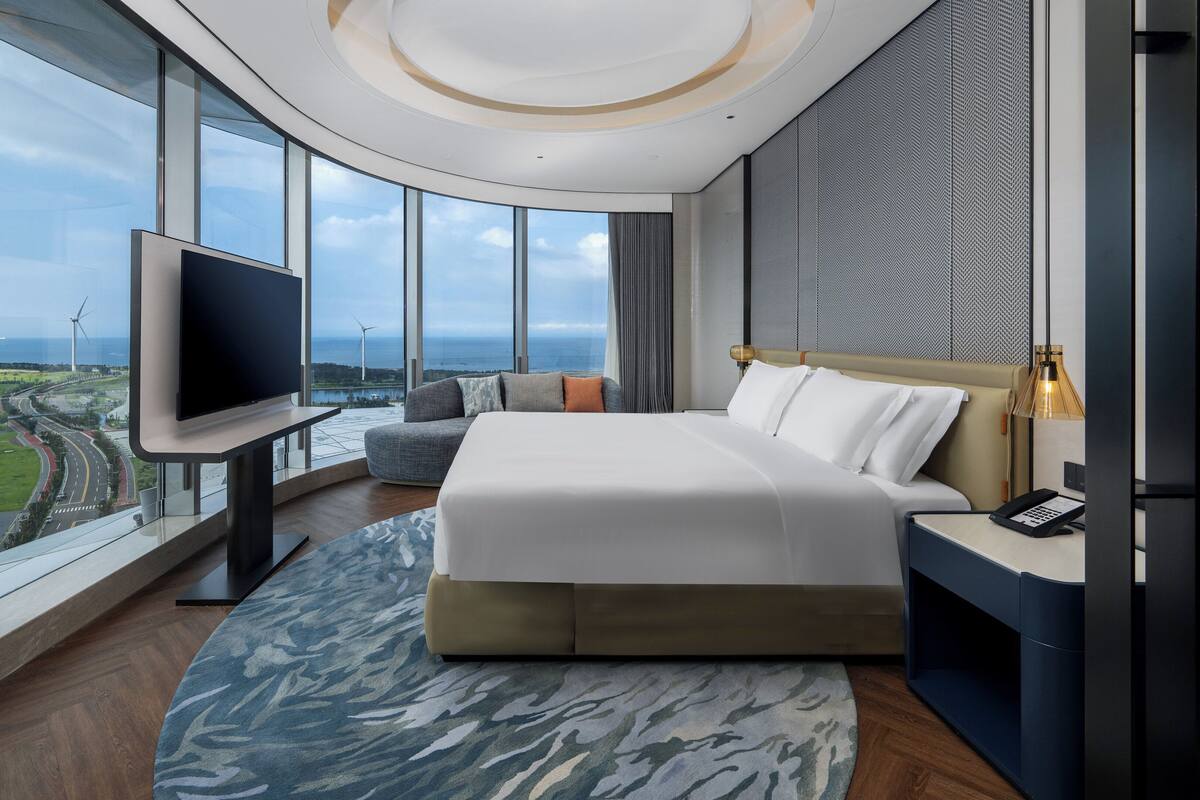 A modern hotel room with a large bed, a flat-screen TV, and a view of a cityscape and wind turbines.